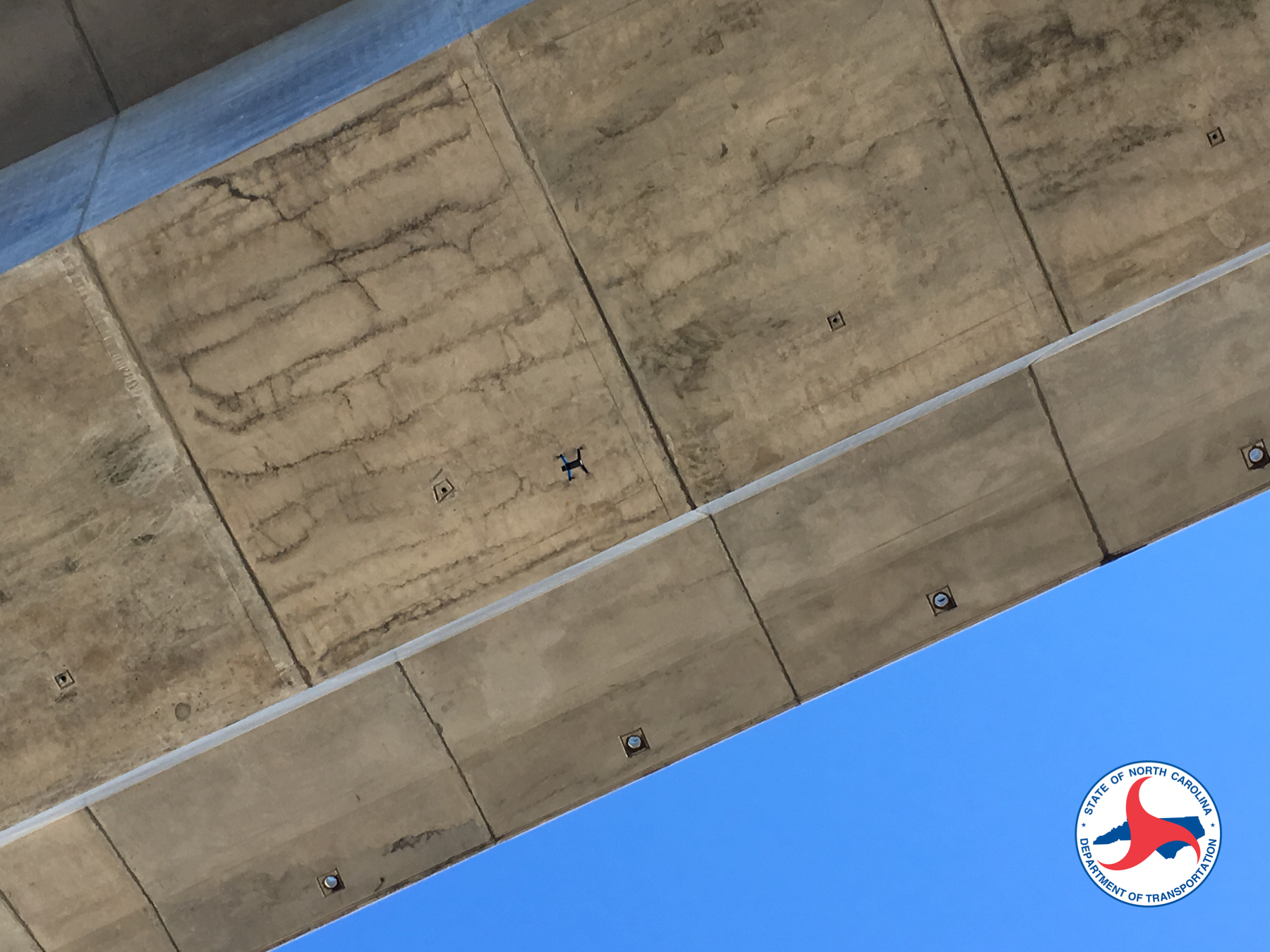 A small blue and black quadcopter hovering close beneath the underside of a concrete bridge deck, photographed looking upward from below. Weathered concrete panels with staining and drainage fittings fill most of the frame, with a wedge of blue sky at the lower right.