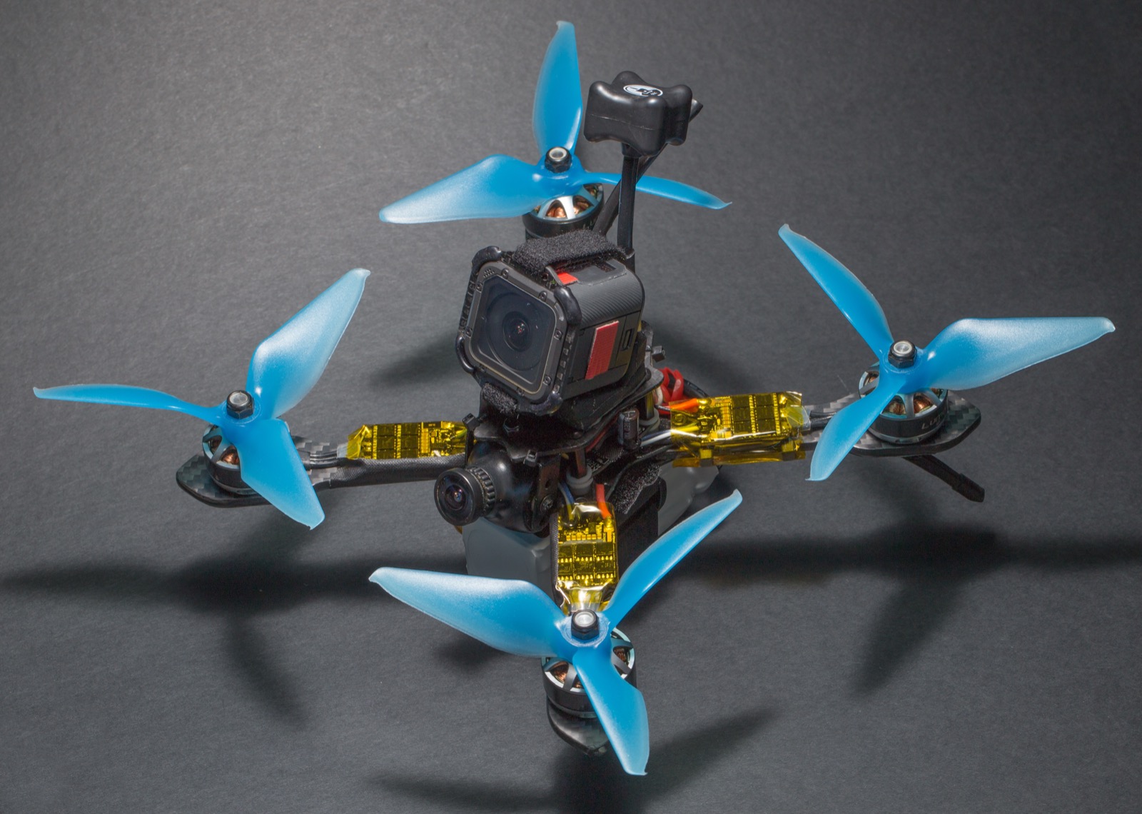 A small carbon-fibre quadcopter frame photographed from above at an angle, with four brushless motors mounted at the ends of its arms, an electronics stack sandwiched between the top and bottom plates, wiring running along the arms, and a cloverleaf video transmitter antenna projecting upward.